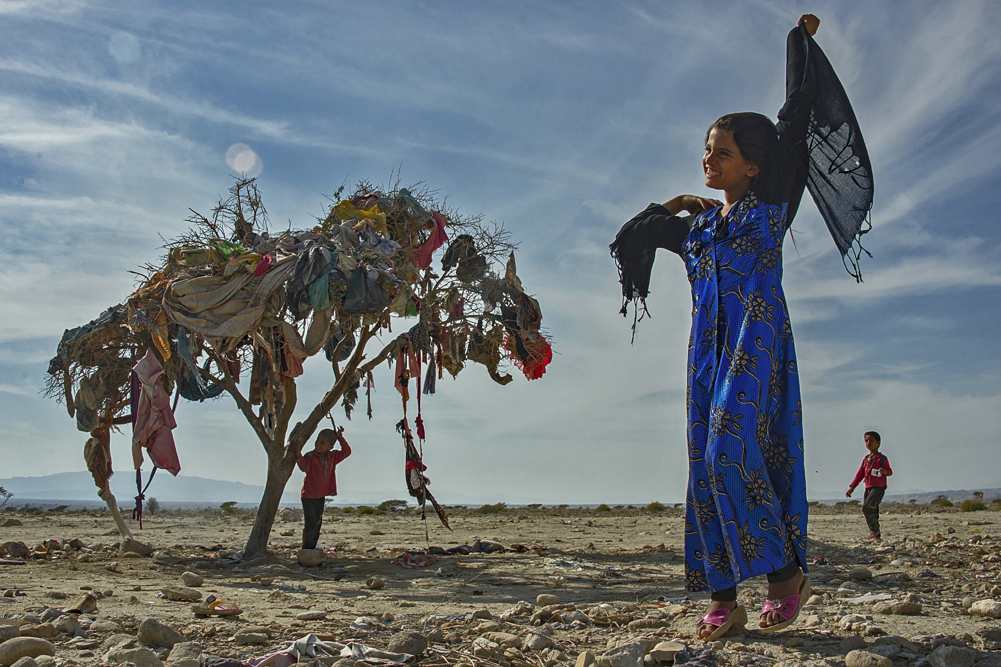 A smiling young girl, lifts her scarf overhead on a clear, sunny day. In the background, a young bow stands underneath a tree covered in debris.