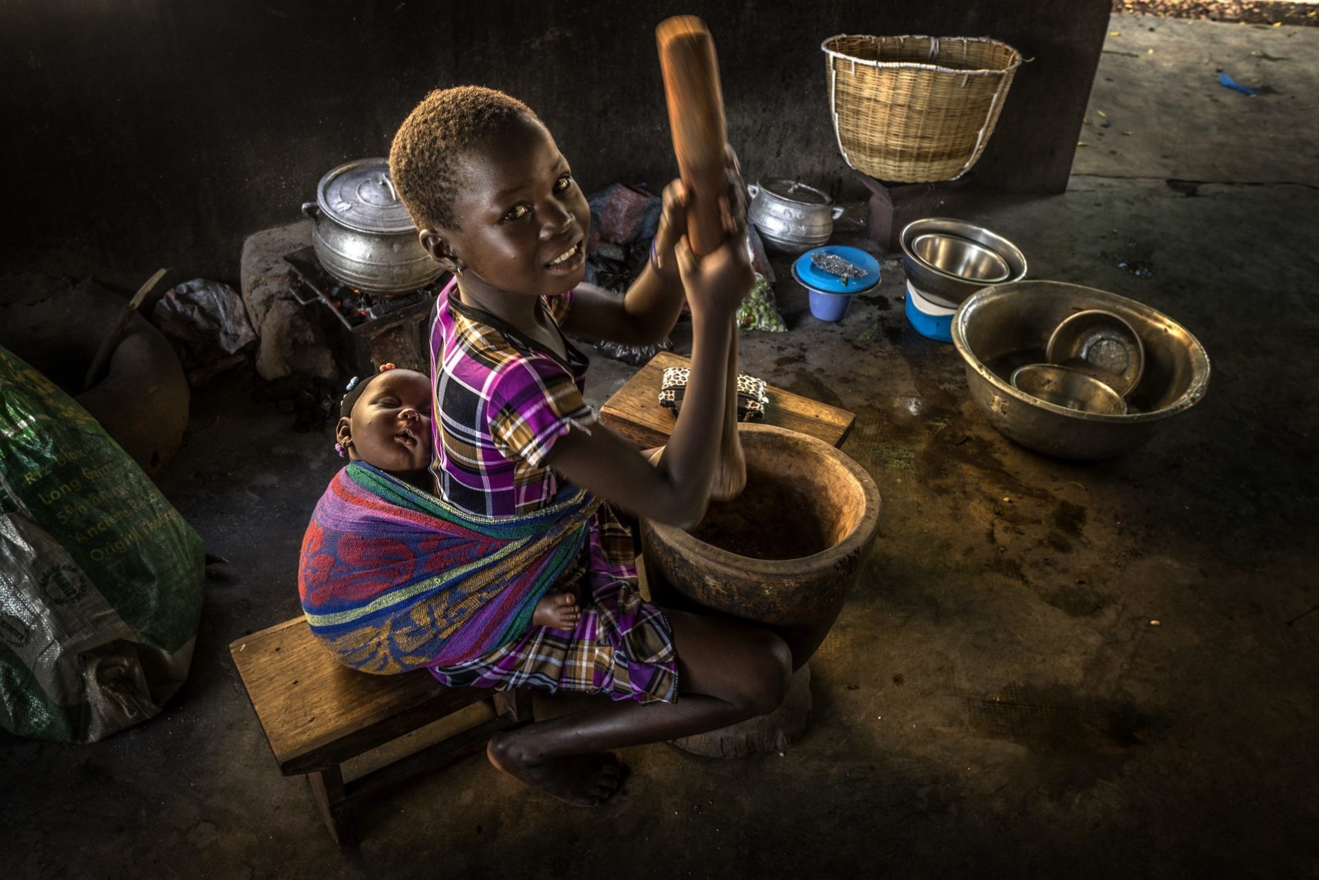 A young smiling west african girl, with a baby strapped to her back, prepares a meal in a large bowl with a wooden masher.