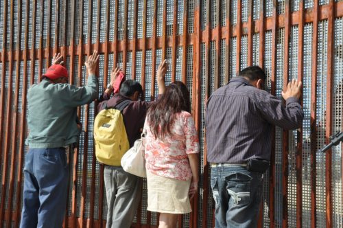 Three men and a woman facing large metal structure separating the U.S. and Mexico