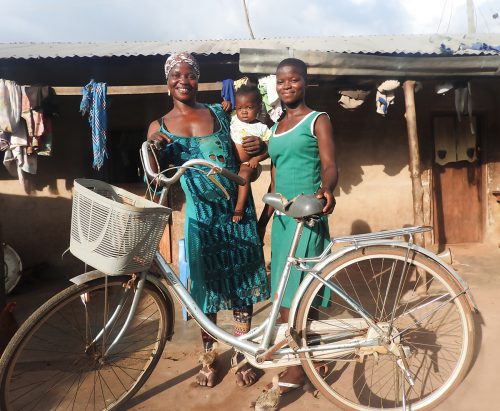 Two women stand in behind a bike, smiling as they pose for the camera. One woman holds a child in her hands.
