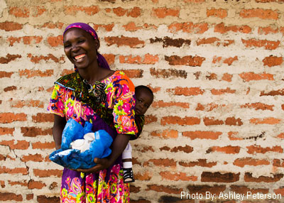 Women, smiling, carries mushrooms in a basket, and a small child on her back. She poses for the camera with a brick wall as the backdrop.