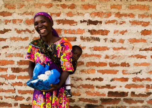 Woman smiles for the camera, as she holds a basket full of mushrooms in front of her, and a young child on her back. She stands in front of a brick wall.