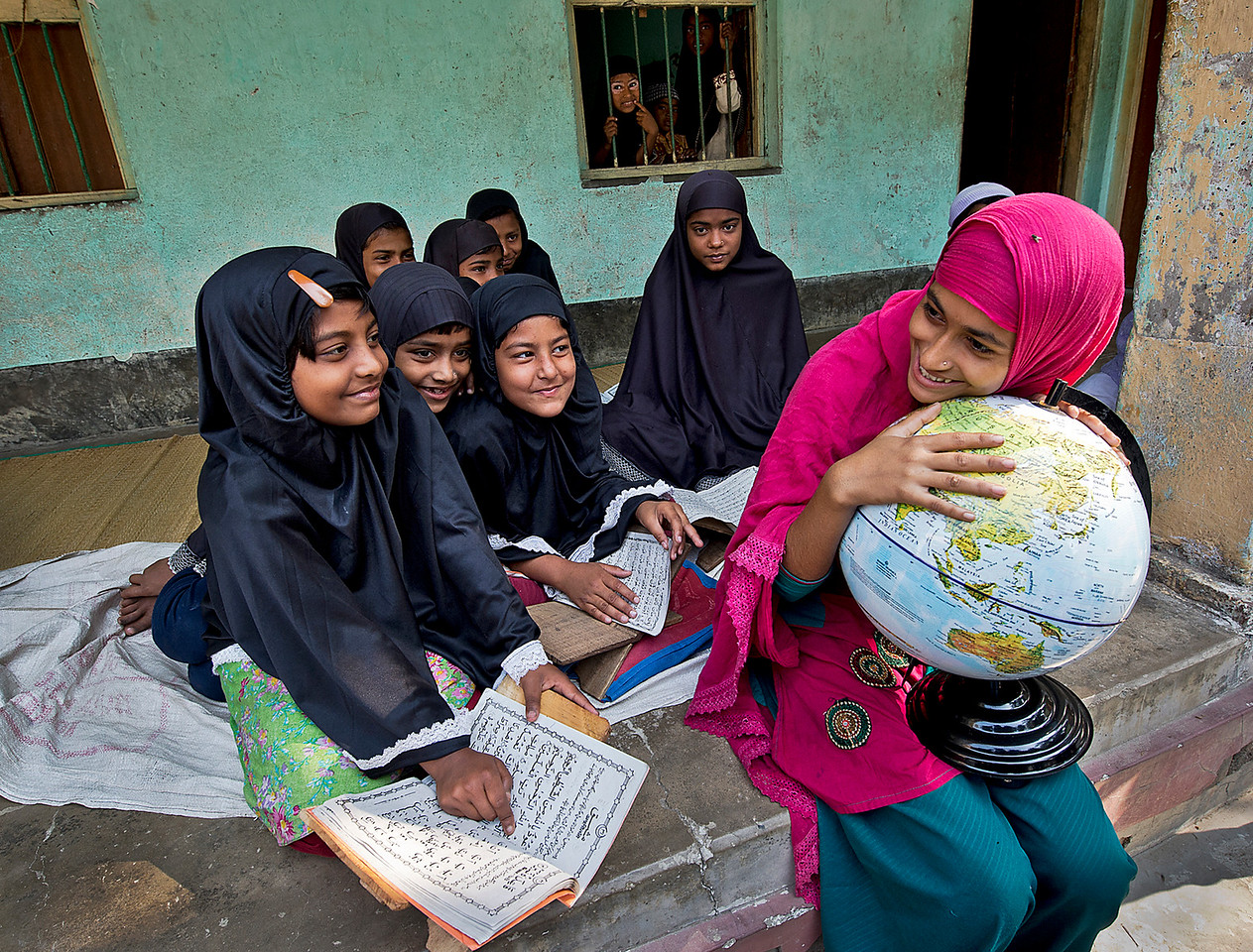 Young girls sit outside of classroom on the landing. The smile as they are learning. While all the girls are dressed in black, one girl stands out in pink, as she holds a globe.