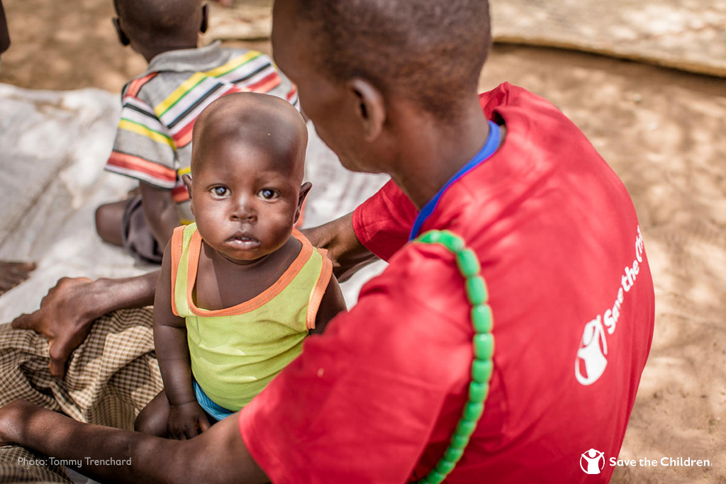 A young child sits in the lap of a humanitarian worker wearing a Save the Children t-shirt