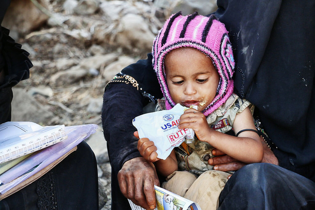 A small child eats a nutritional supplement meal provided by USAID in Yemen, while her mom holds her.