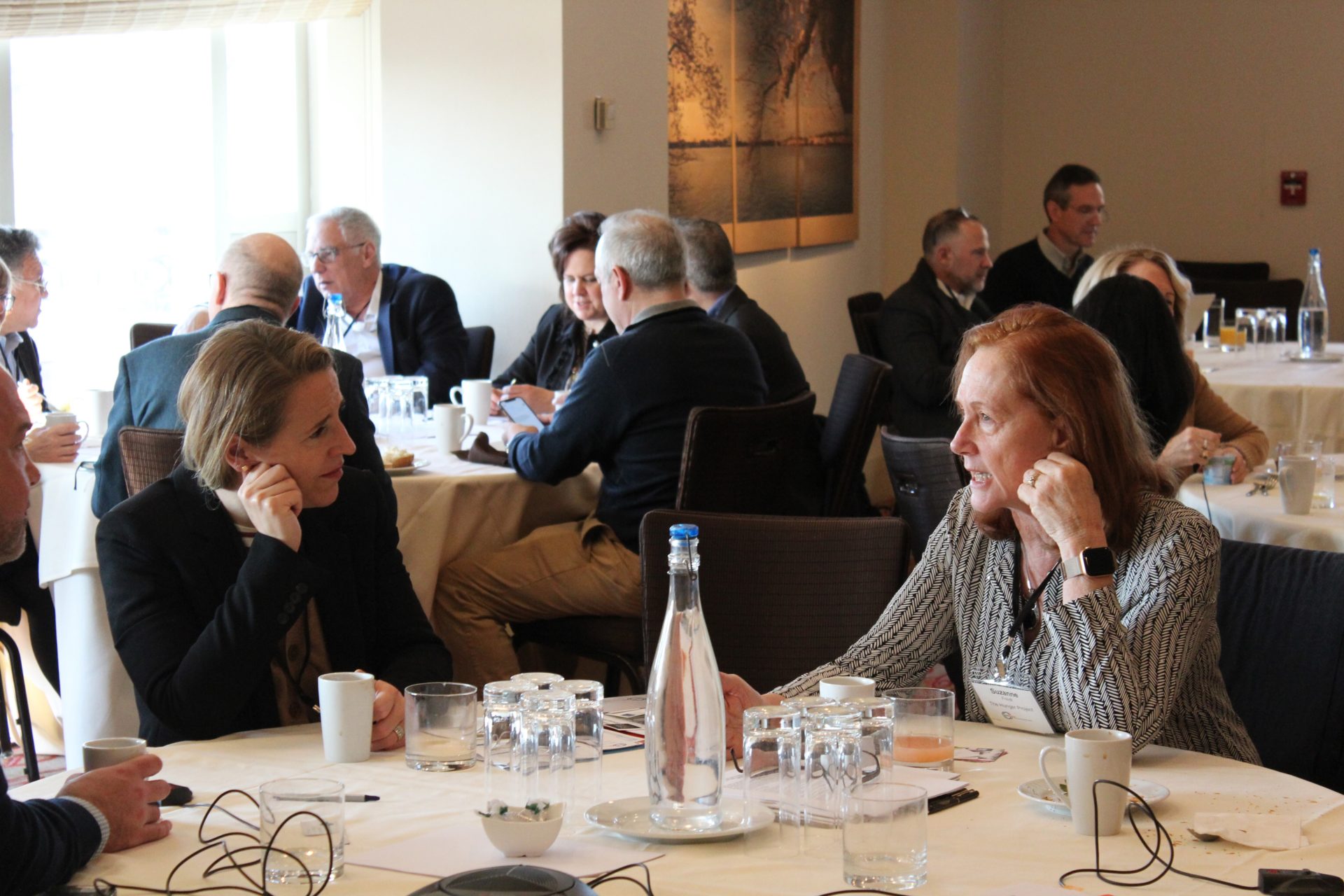 people sit around various tables during a meeting. In the foreground, two women speak at a table.