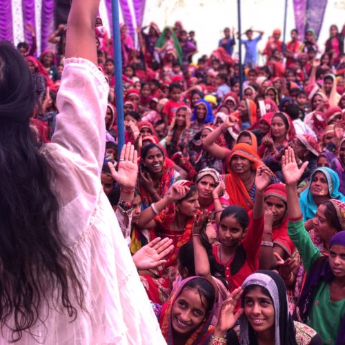 Women Stands in front of crowd, with her back to the camera, leading them as they raise their hand in agreement. The women are dressed in colorful garbs and smiling.