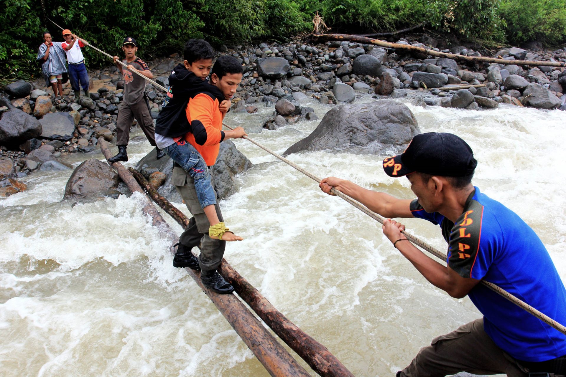 Using a rope, a man helps pull several men and boys cross a river on two thin logs.