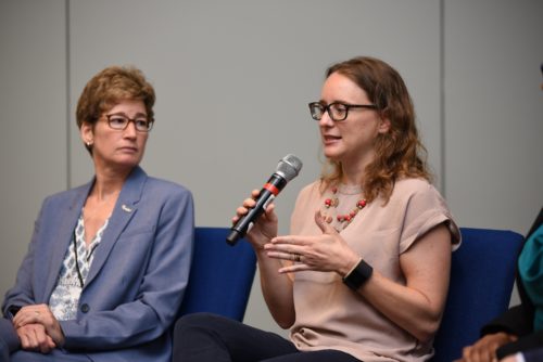 two women sit on stage. One holds a microphone to speak as the other looks on.