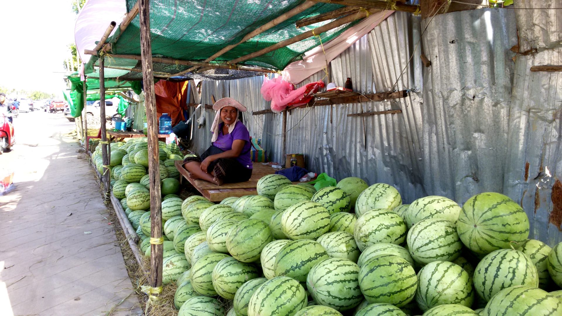 Young women sits under her stand where she sells a large number of watermelons.
