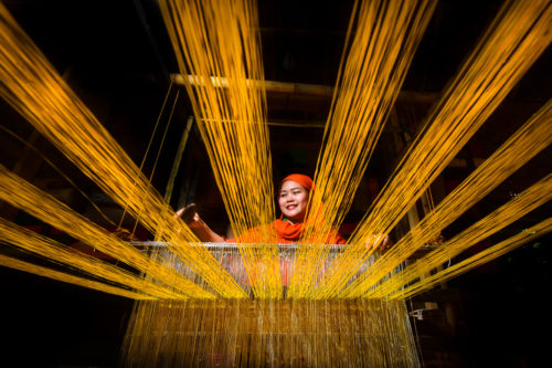A women stands behind a loom weaving yellow thread