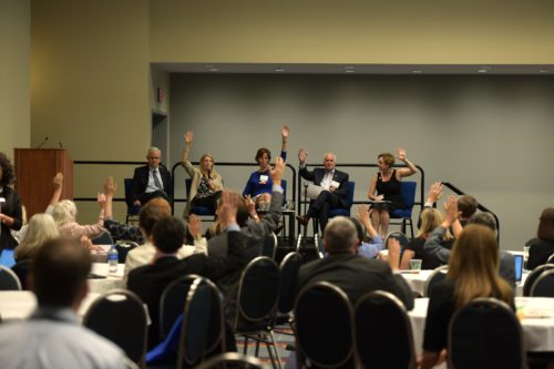 people in a room sitting at round tables and on a stage. Someone asks a squestion and those who agree raise their hands.