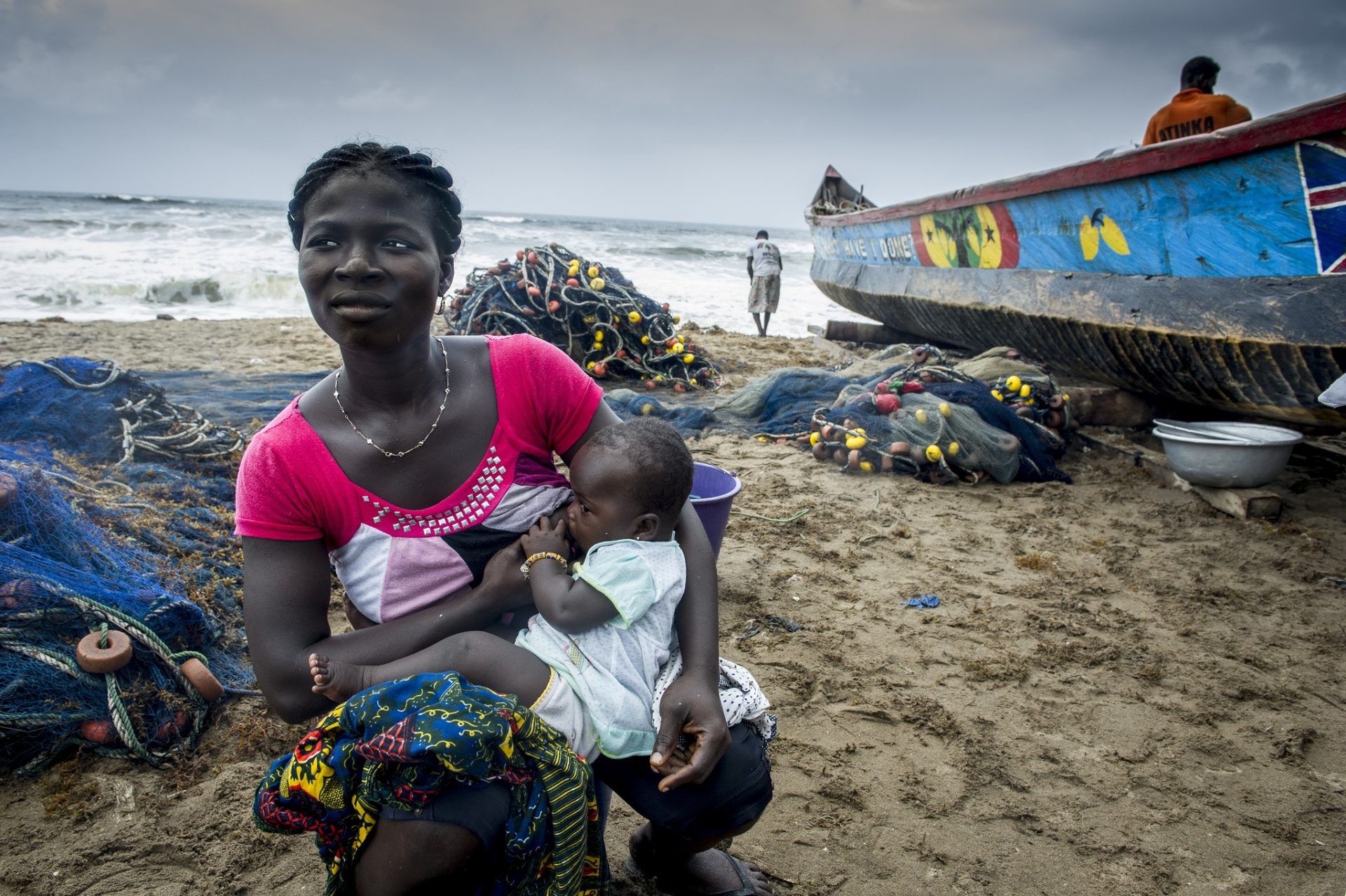 Women holds baby on the beach shore. Boat washed up ashore in the background.