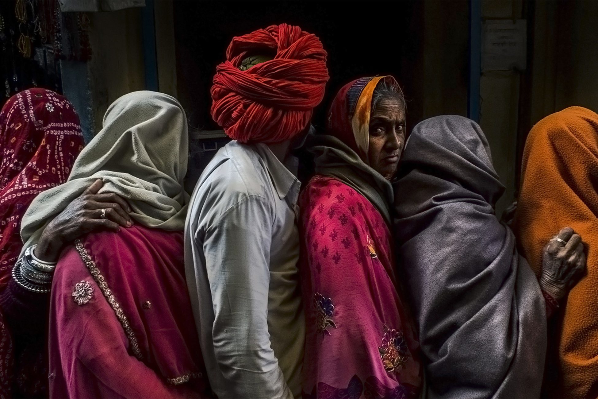 a group of men in women stand in line. All them are fully covered, faces hidden, with the exception of one woman who looks boldly at the camera.