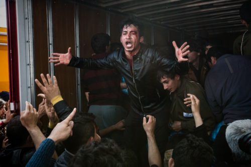 A man stands inside the back of a truck yelling at an eager crowd as they approach the truck looking for help.