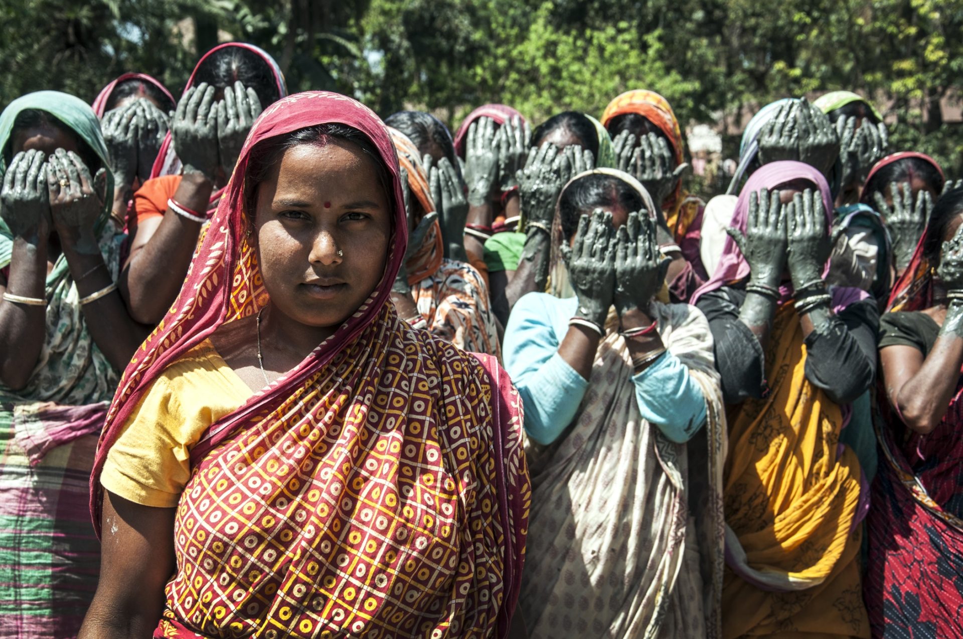 A woman stands at the forefront, staring directly into the camera. A group of women stand behind her cover their faces in protest.