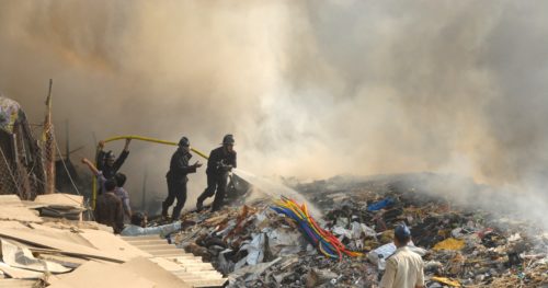 Firemen spray water on a large pile of smoldering rubble. Smoke fills the sky behind them.