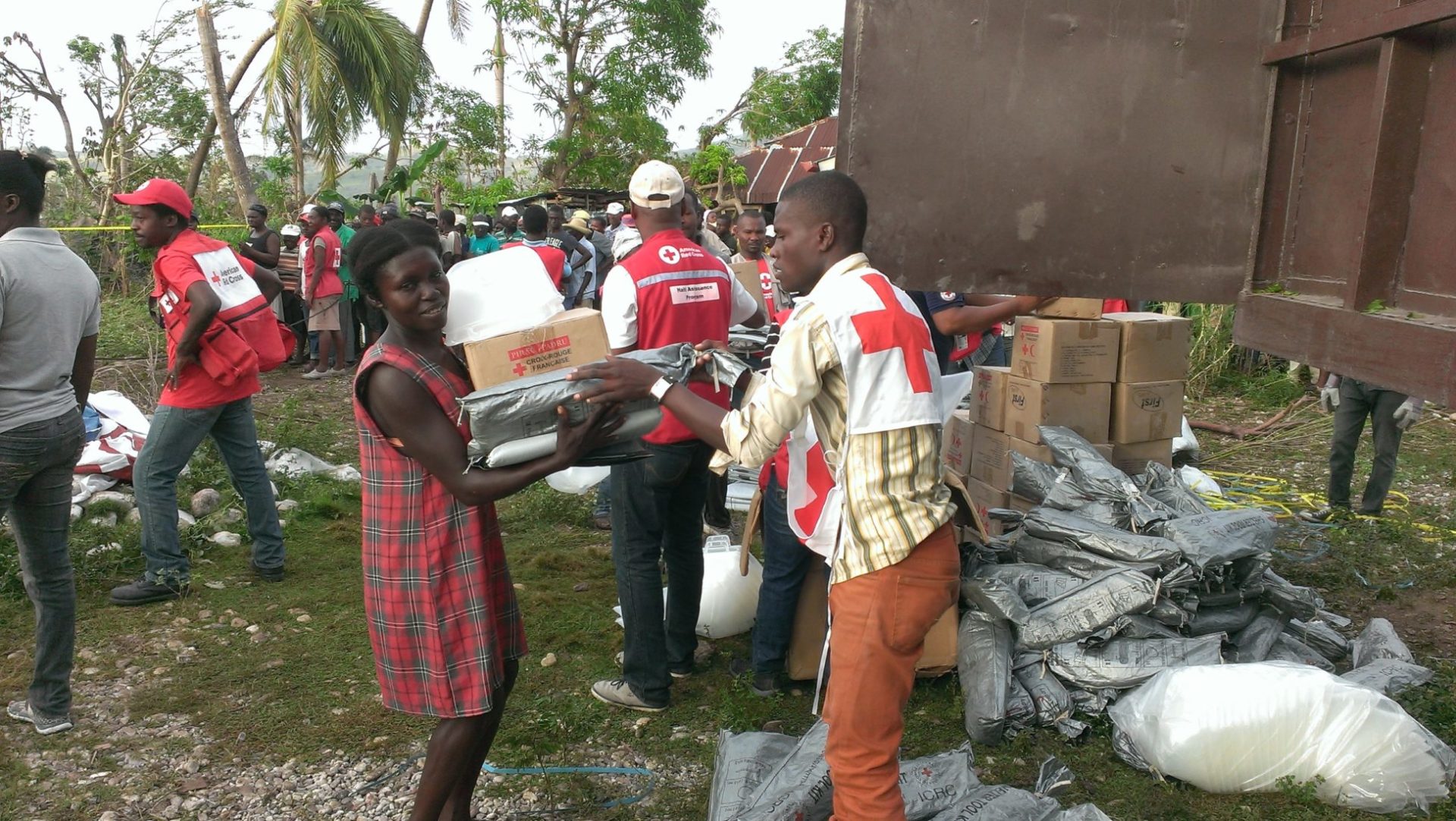 Red Cross aid worker hands over a number of items to a young woman. In the background, other items are distributed to a waiting crowd.