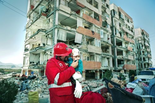 A rescue worker hold a small injured child in front of a partially destroyed apartment building