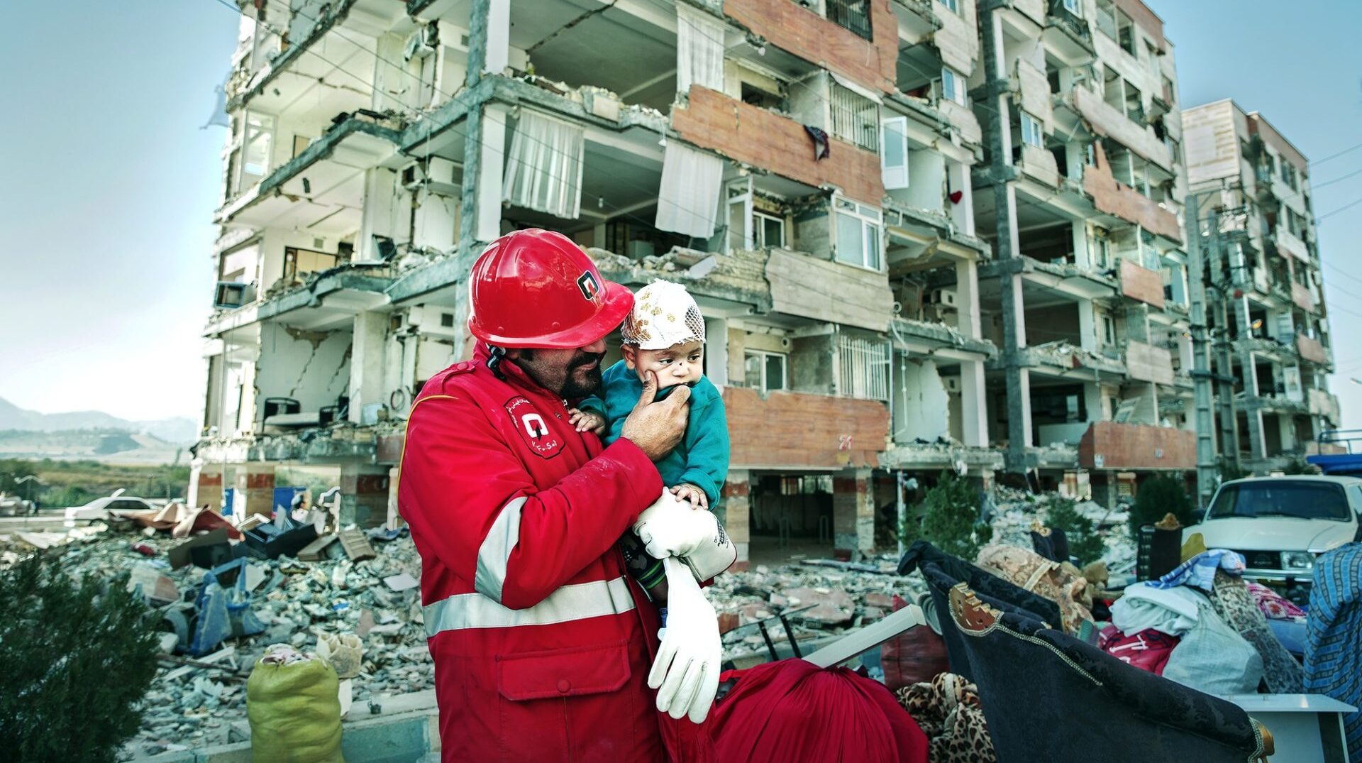 A rescue worker hold a small injured child in front of a partially destroyed apartment building