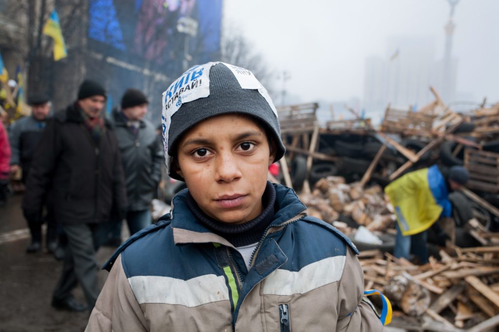 In the midst of a rubble, a young and determined looking boy, wearing a coat and warm hat, poses for the camera