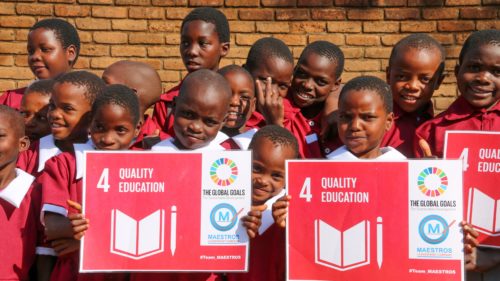 A group of school - children dressed in red school uniforms - pose for the camera, holding up placards with sustainable development goal number 4.