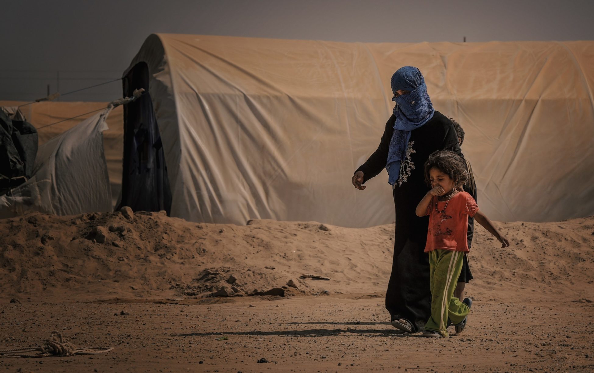 A woman and her young child walk in the middle of the desert passing by a large makeshift shelter for displaced people.