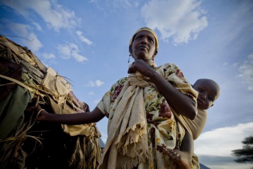 A woman poses for photographs with, a young child strapped to her back, at her home in Kenya. Backdrop is a blue sky and a tent like structure where the woman lives. Photo taken from below.