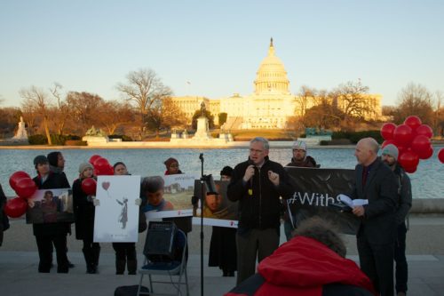protesters stand across the river from capitol building, holding signs reading #withsyria