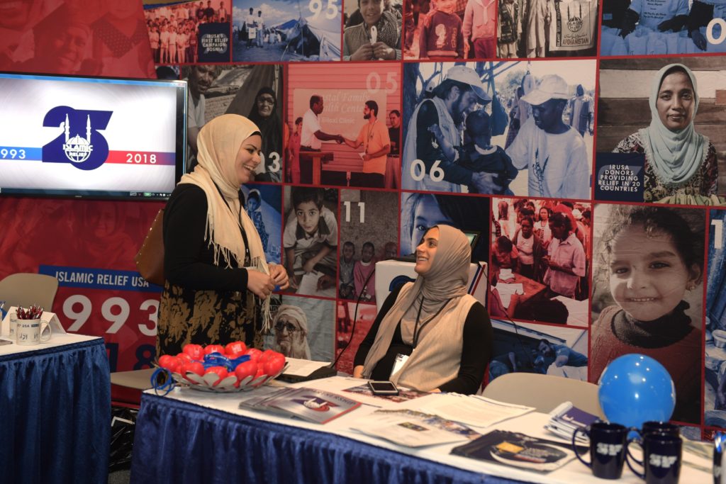 two women, one standing, on sitting, facing one another at the booth covered in color images from Islamic Relief.