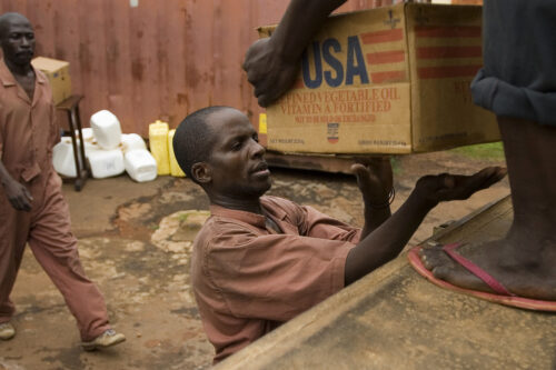 A man in work clothes prepares to take a USAID-branded cardboard box of vegetable oil from another person off-camera who is standing above him on the back of a truck.