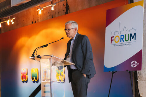 Sam Worthington stands on stage at InterAction's 2022 Forum in front of a colorful art gallery wall and a Forum sign.